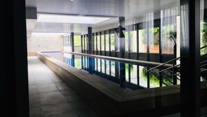 Una piscina interior moderna con paredes de vidrio transparente, suelo de baldosas y una escalera que lleva a un nivel superior.