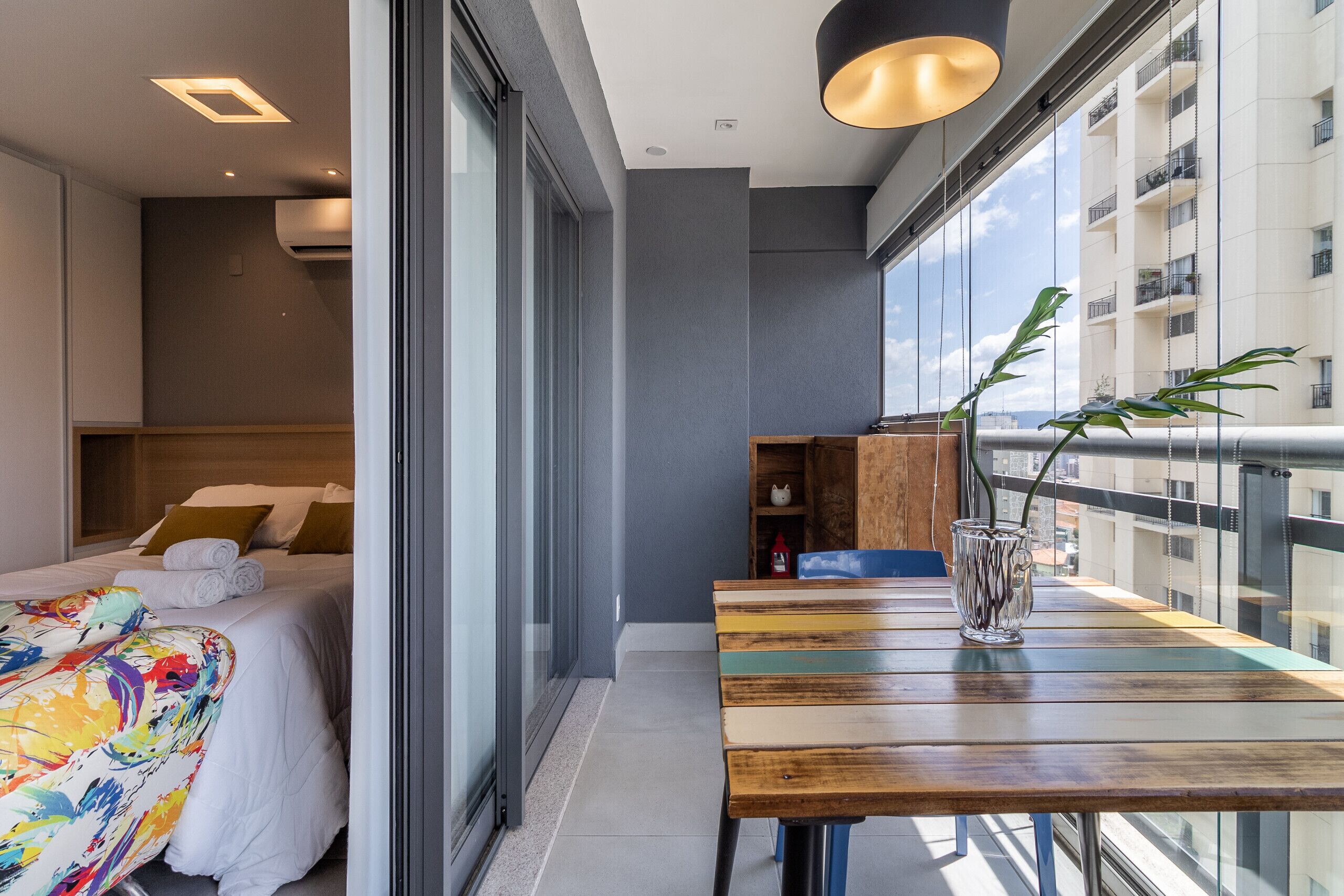 A modern hotel room with a large bed, a wooden table with a vase, and a balcony with a view of the city.