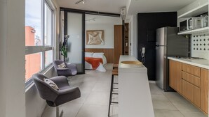 Cocina moderna con refrigerador, microondas y gabinetes de madera. Hay un comedor con mesa y sillas, y una cama con manta roja.