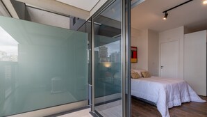 Een moderne slaapkamer met een glazen deur naar een balkon, een bed met wit beddengoed en uitzicht over de stad.