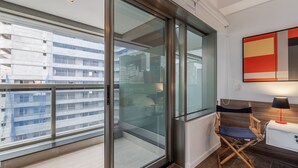 Een moderne hotelkamer met een grote schuifglazen deur naar een balkon, een regisseursstoel, een klein wit nachtkastje en een abstract schilderij in rood en beige op de muur.