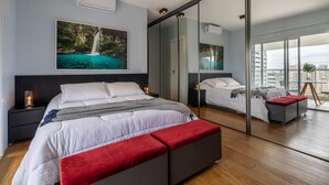 A modern bedroom with a large bed, a red bench, a nightstand with a lamp, a wall-mounted air conditioner, and a large window with a city view. - Baixo Augusta queen bed, pool, gym and garage (São Paulo)