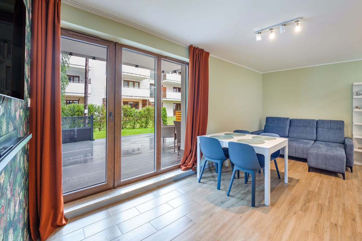 Ein modernes Wohnzimmer mit Esstisch, blauen Stühlen, einem grauen Sofa und einer Glastür zum Balkon.