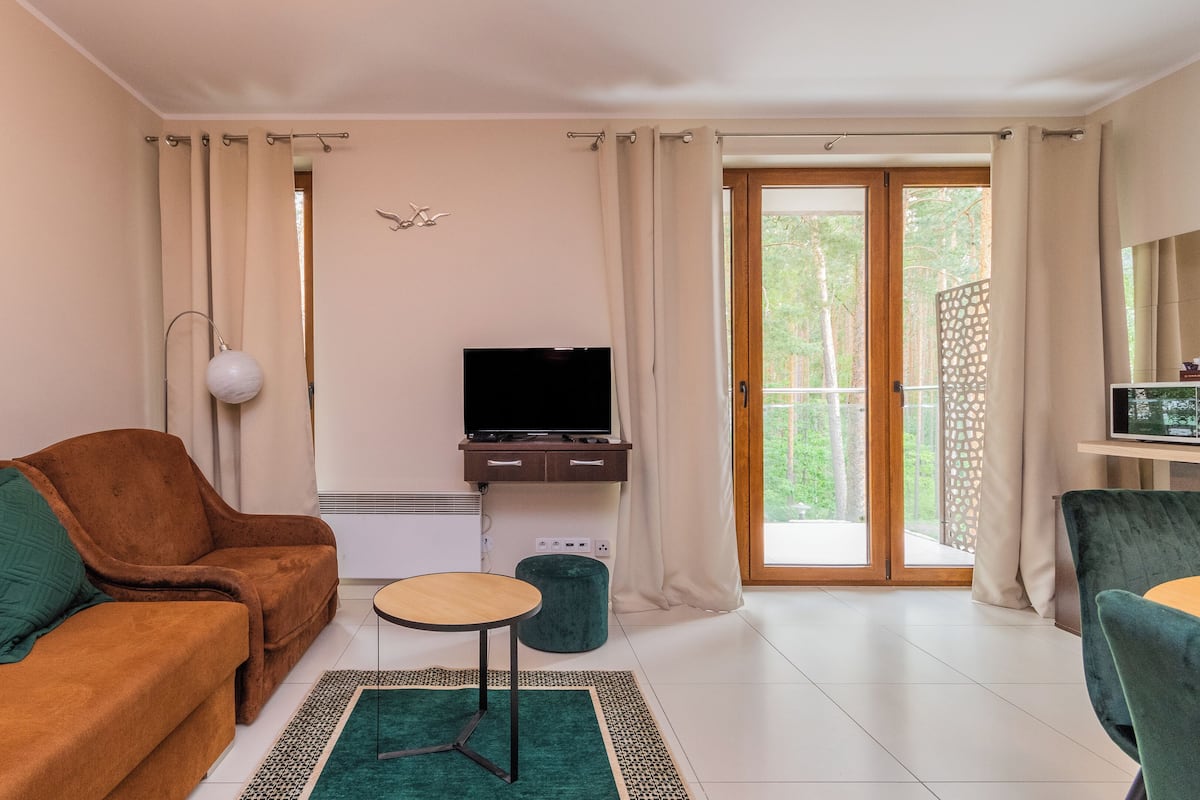 Ein Wohnzimmer mit braunem Sofa, einem kleinen runden Tisch, einem Fernseher auf einer Holzkommode und Blick auf einen Wald durch eine Glastür.