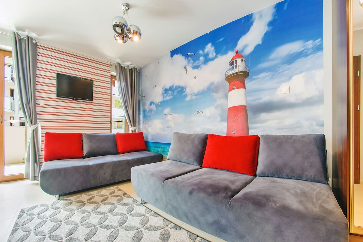Ein Wohnzimmer mit einer großformatigen Wandmalerei eines Leuchtturms, grauen Sofas mit roten Kissen, einem wandmontierten Flachbildfernseher und einem gemusterten Teppich.