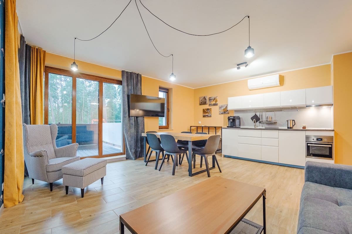 Eine moderne Küche mit weißen Schränken, einem eingebauten Ofen und einer Kaffeemaschine. Es gibt eine Essecke mit Tisch und Stühlen, und ein Sofa im Wohnbereich.