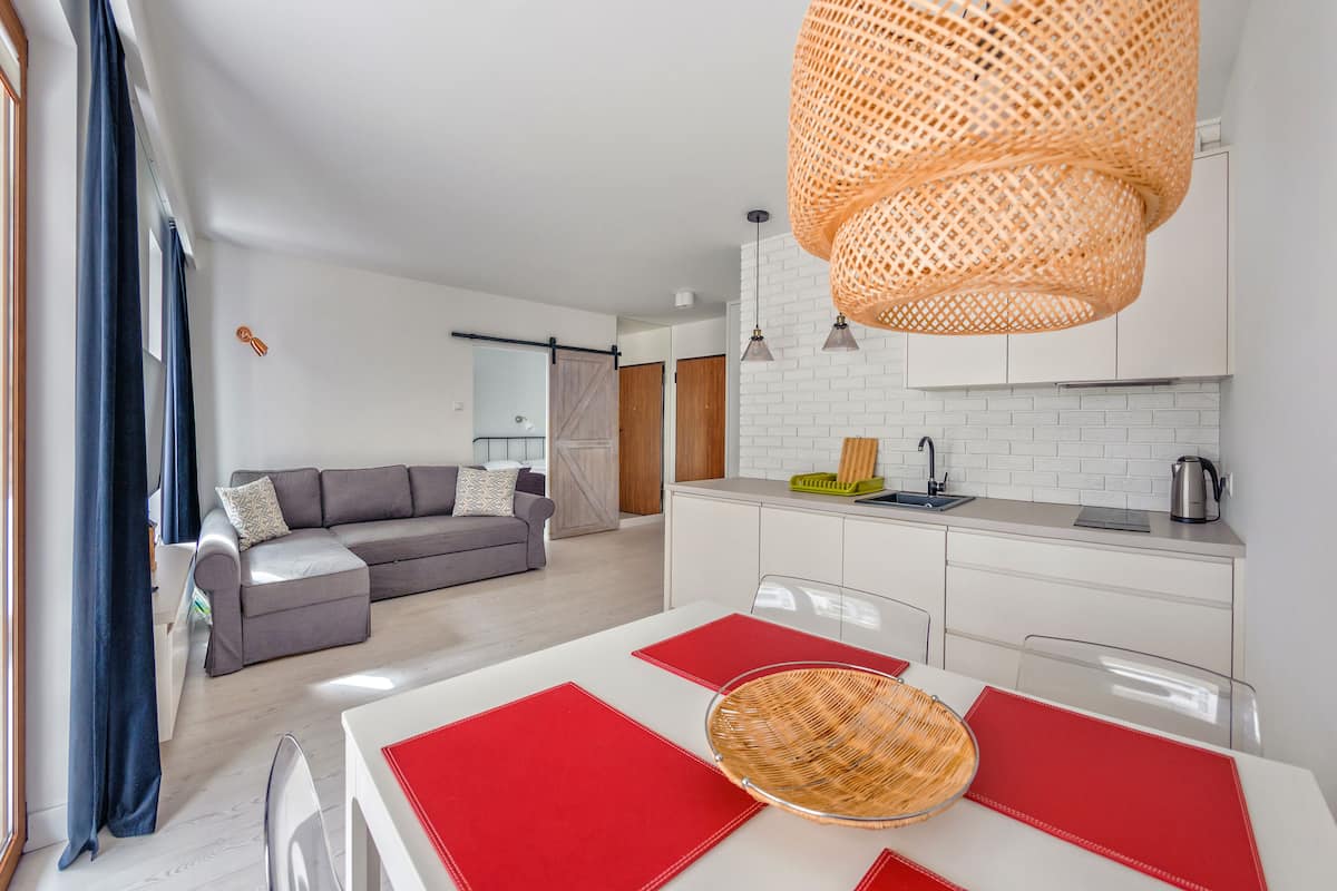 Ein modernes Wohnzimmer mit einem grauen Sofa, einem Esstisch mit roten Platzsets und einer Küchenzeile mit weißen Schränken und einer hängenden, orangefarbenen Leuchte.