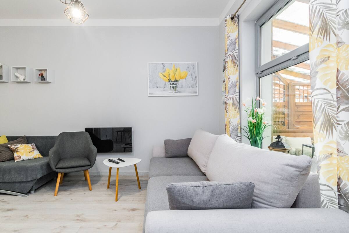 Ein modernes Wohnzimmer mit grauem Sofa, grauem Sessel und weißem Couchtisch. An der Wand hängen drei gerahmte Bilder, ein Fenster mit Vorhängen und eine Pflanze ist rechts zu sehen.