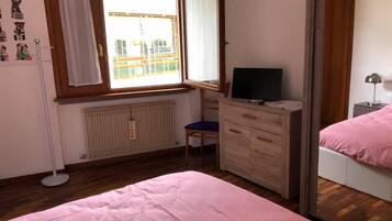 A bedroom with a bed, a dresser, a chair, a radiator, a window with curtains, a mirror, and a TV.