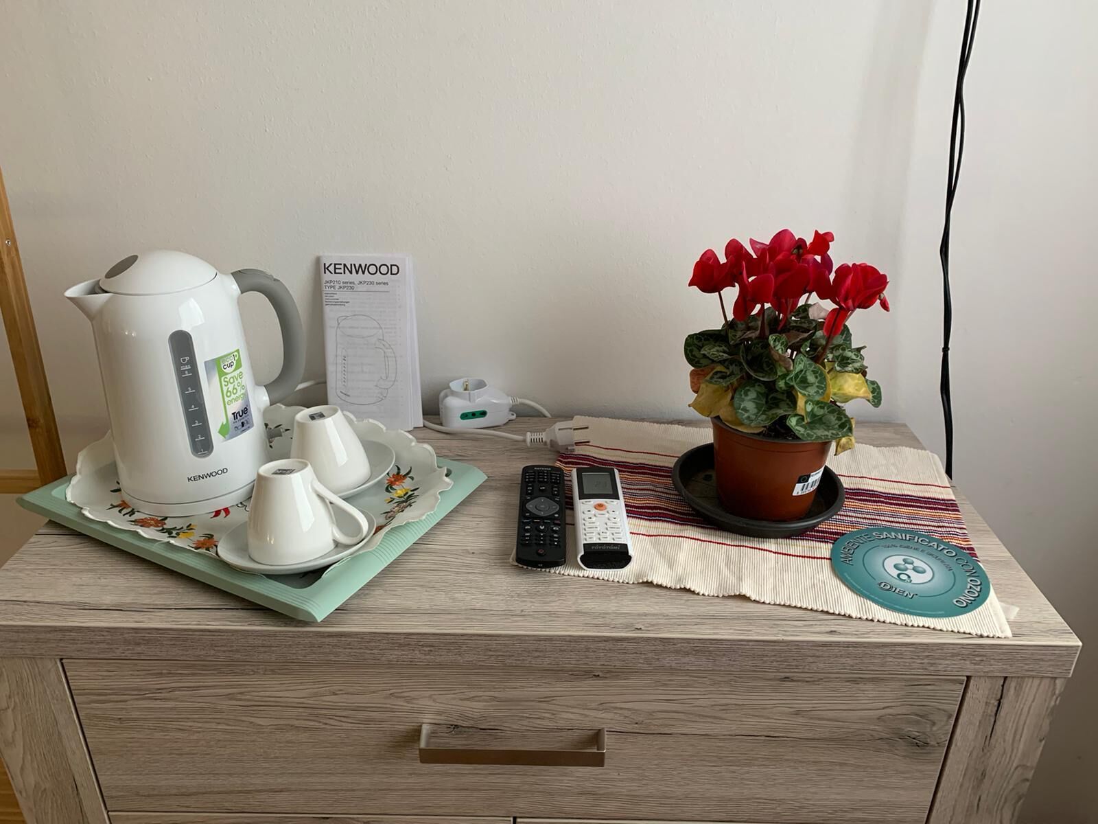 Una mesita de noche de madera con una tetera, tazas de té, un control remoto, un teléfono inteligente, una planta en maceta y un mantel individual.