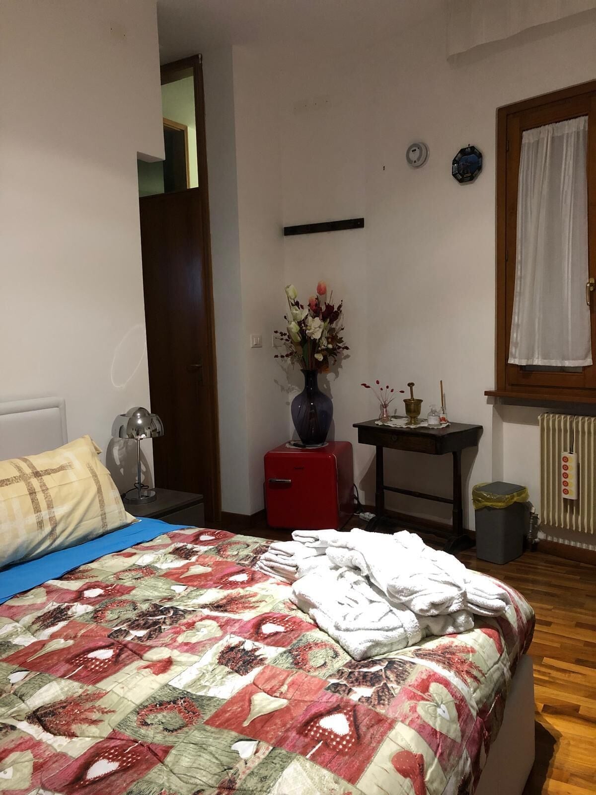 Un dormitorio con cama, mesita de noche, aparador rojo, florero con flores, pequeña mesa con lámpara y ventana con cortinas.