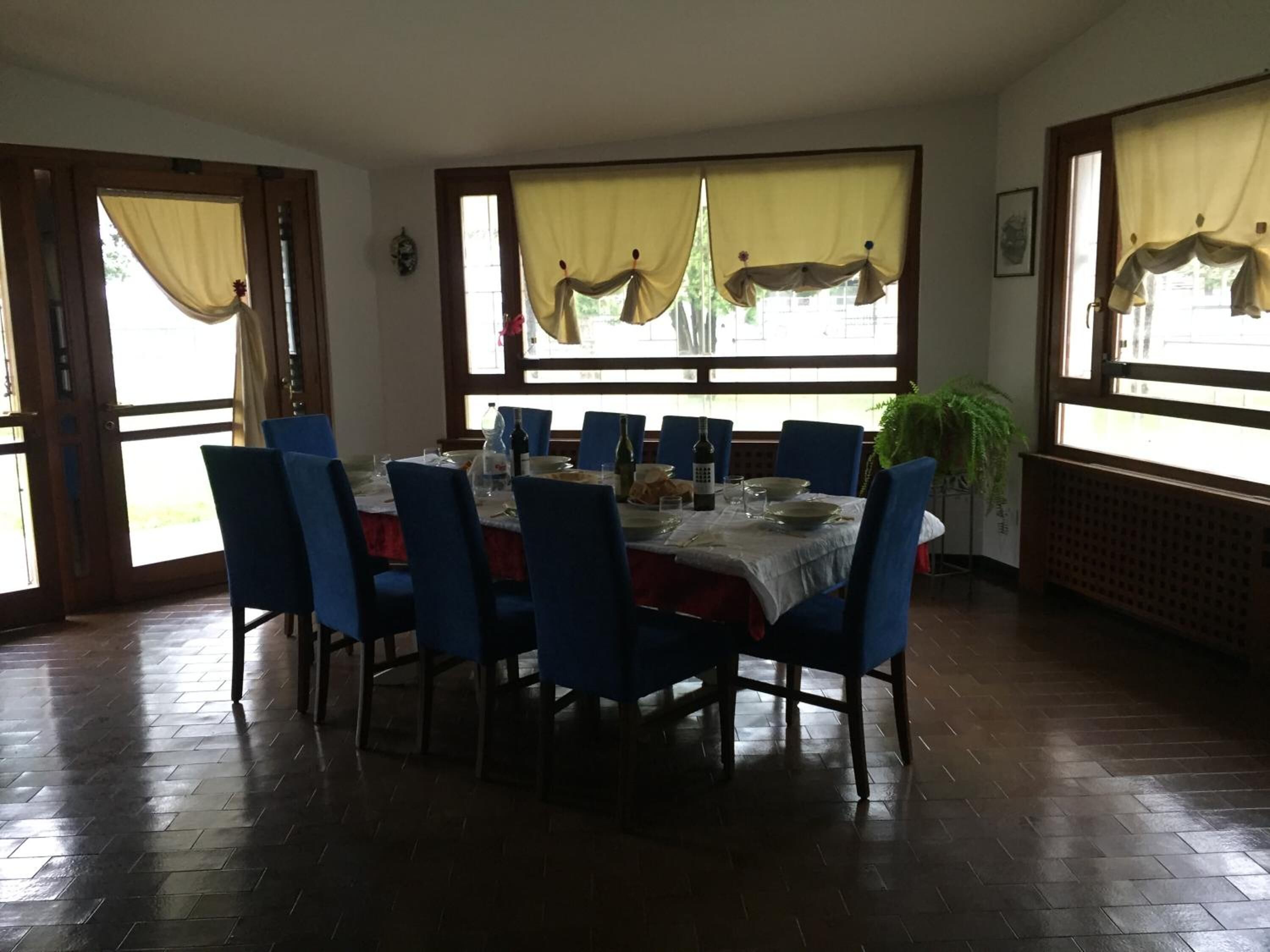 Zona pranzo con tavolo rotondo apparecchiato per sei persone, circondato da sedie blu. Alle finestre sono presenti delle tende e in un angolo si nota una pianta in vaso.