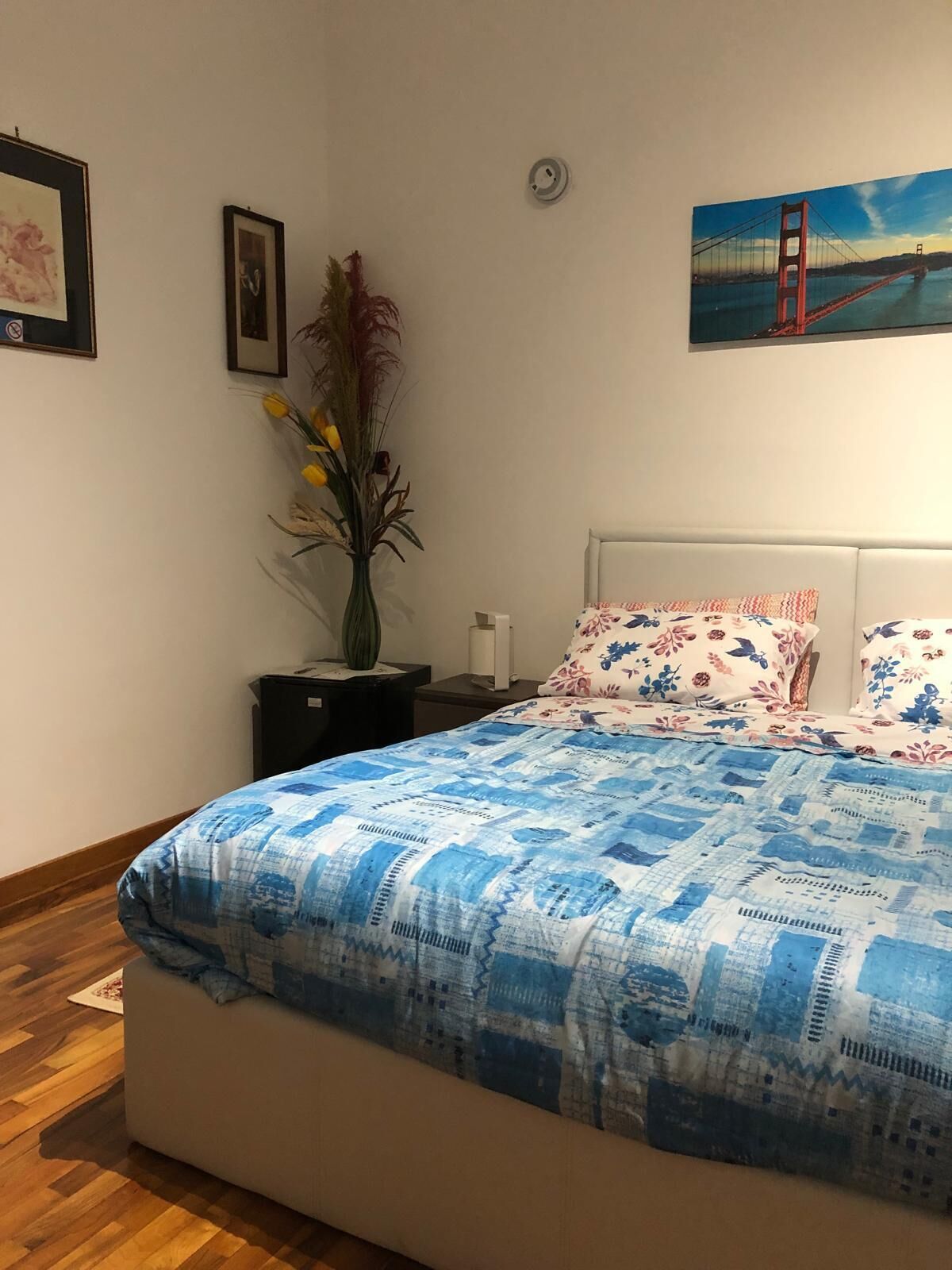 A bedroom with a bed, a nightstand, a vase with flowers, a framed picture, and a painting of a bridge.