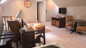 A cozy loft-style room with a high ceiling, a large clock on the wall, a television, a dining table with chairs, and a horse-themed chair.
