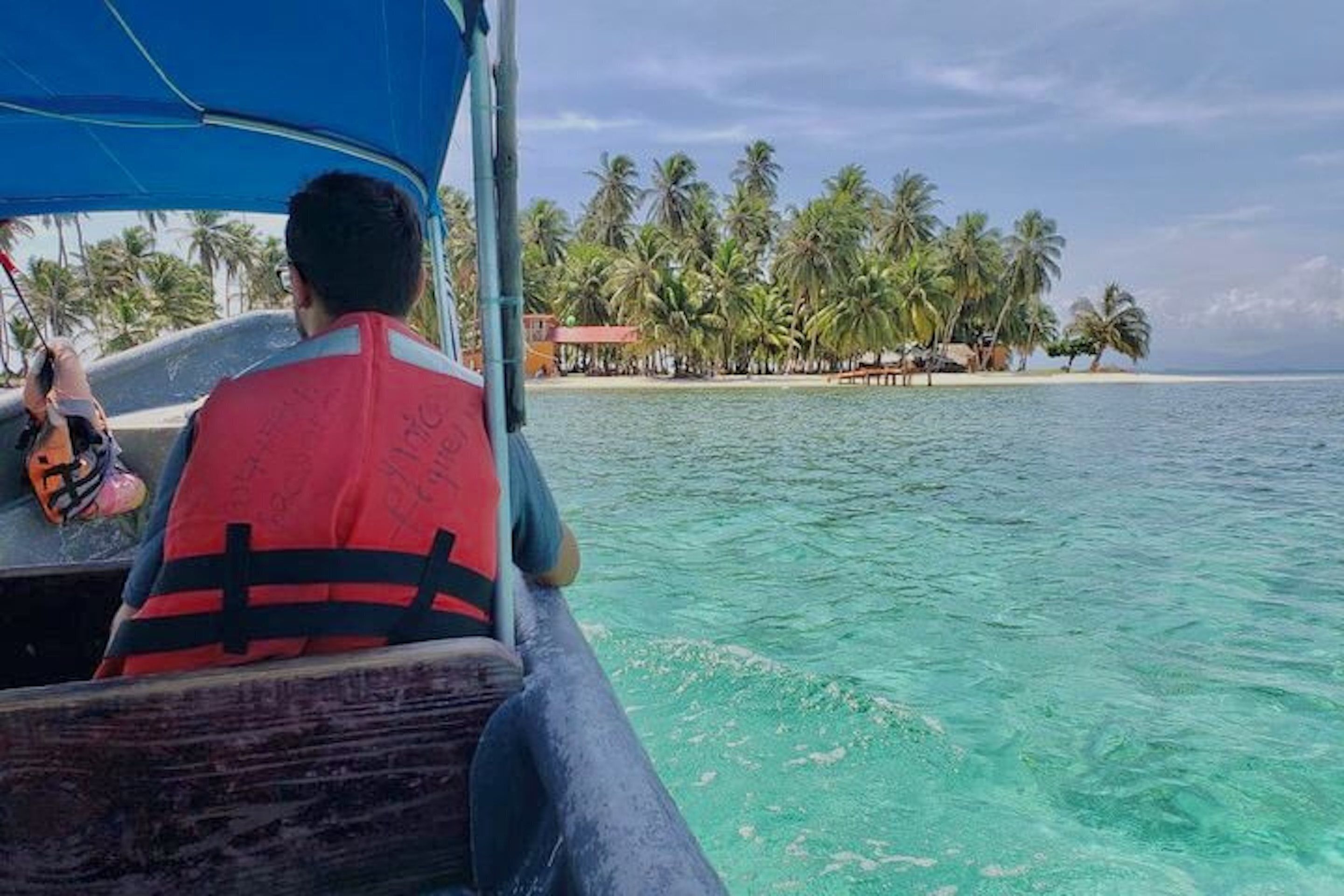Una persona en un bote, con chaleco salvavidas, disfruta de una vista despejada a una playa con palmeras y un pequeño edificio al fondo.