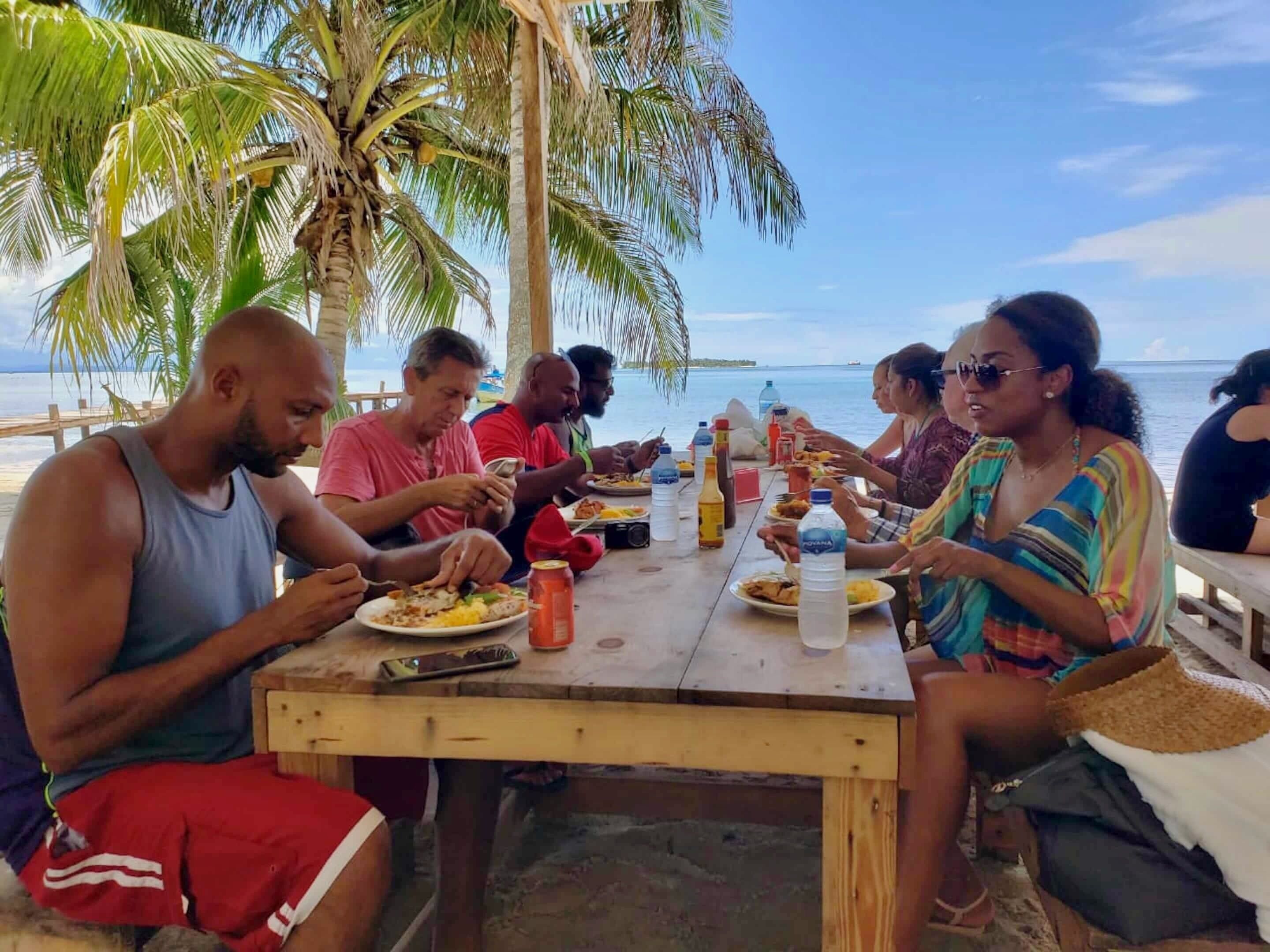 A group of people dining at a wooden table with a view of the ocean and palm trees.