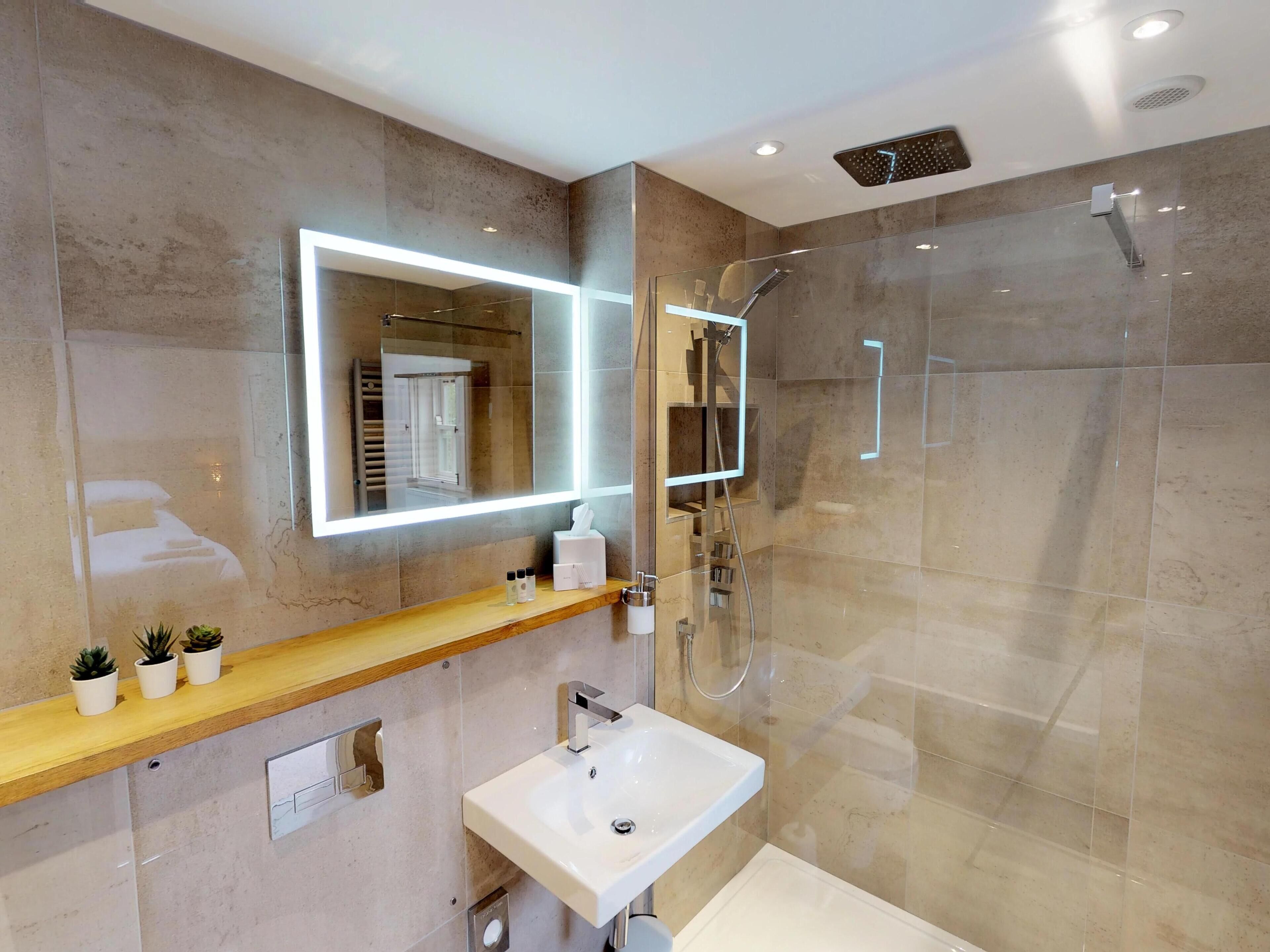 A modern bathroom with a glass-enclosed shower, a large mirror with integrated lighting, a wooden vanity, and a white sink.