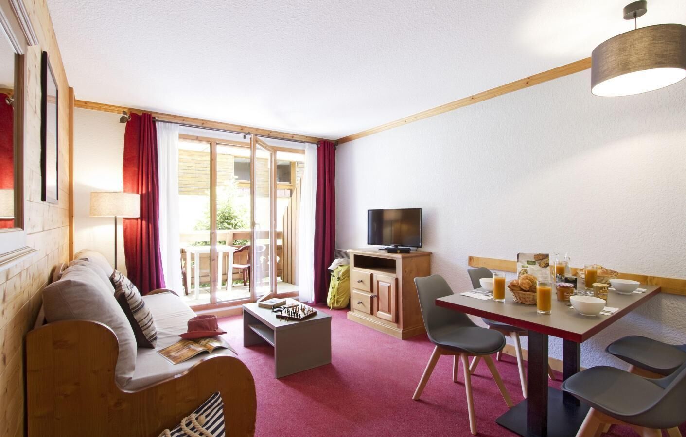 Ein gemütliches Zimmer mit einer Couch, einem Couchtisch, einem Fernseher, einem Esstisch mit Stühlen und einer Schiebetür zu einem Außenbereich.