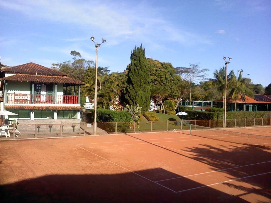 A tennis court with a fence, a building with a red roof, and trees in the background.