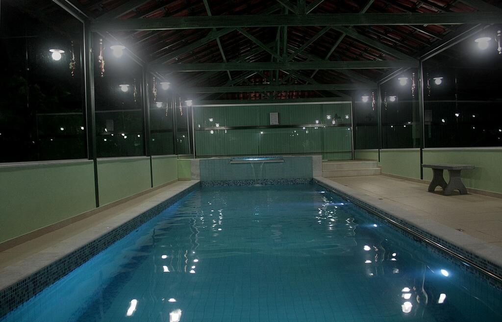 An indoor swimming pool with clear water, surrounded by glass walls and a tiled floor, under a metal roof structure.