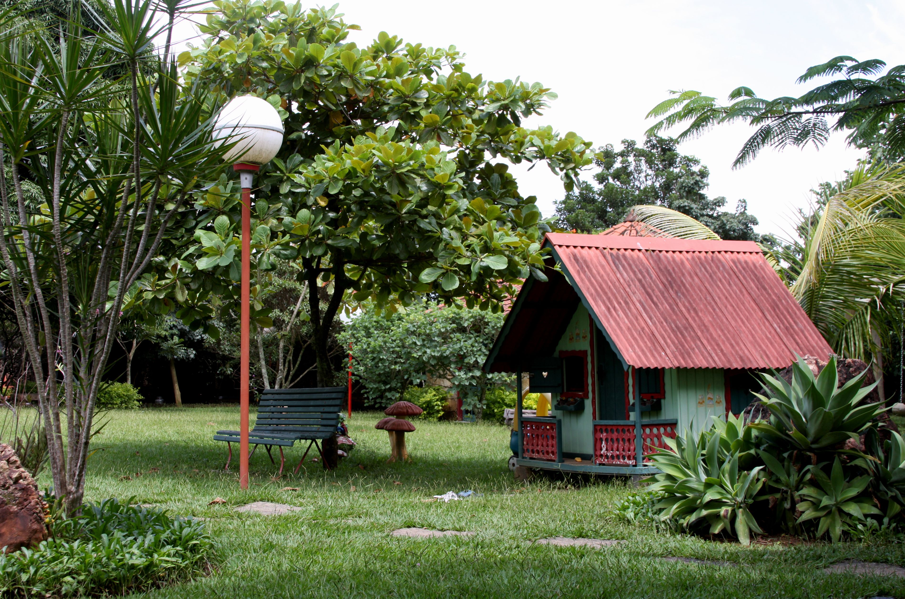 A small, rustic house with a red roof surrounded by lush greenery and a bench.
