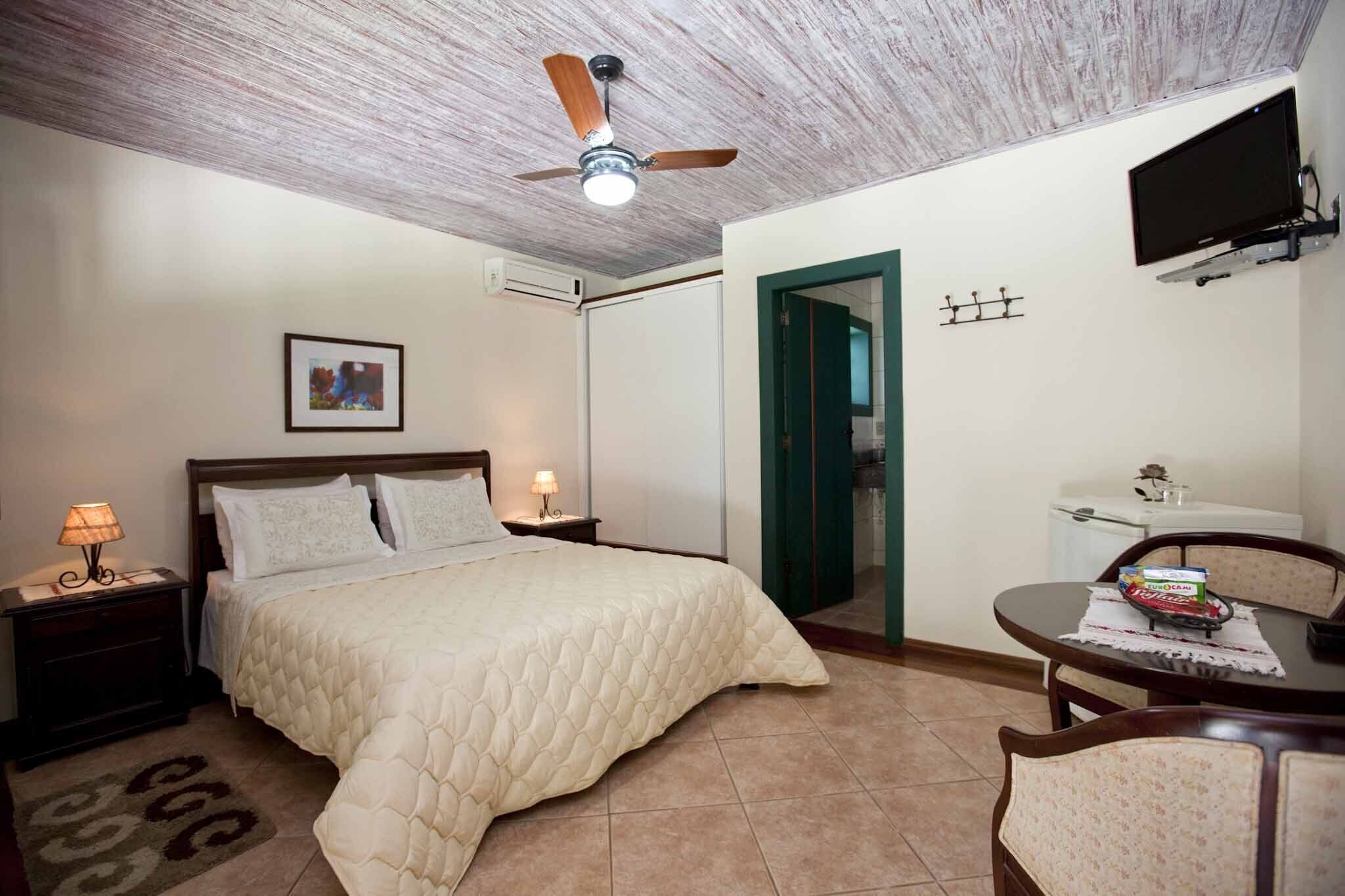 Un dormitorio con cama, lámparas de noche, ventilador de techo, televisor montado en la pared, una pequeña mesa con una silla y una puerta que da a otra habitación.