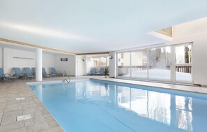 Une piscine intérieure avec des parois en verre transparent, des chaises bleues et un sol carrelé.