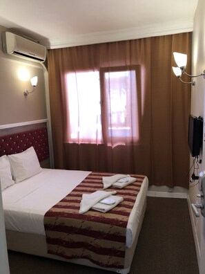 A hotel room with a bed, a flat-screen TV, an air conditioner, and a window with curtains.