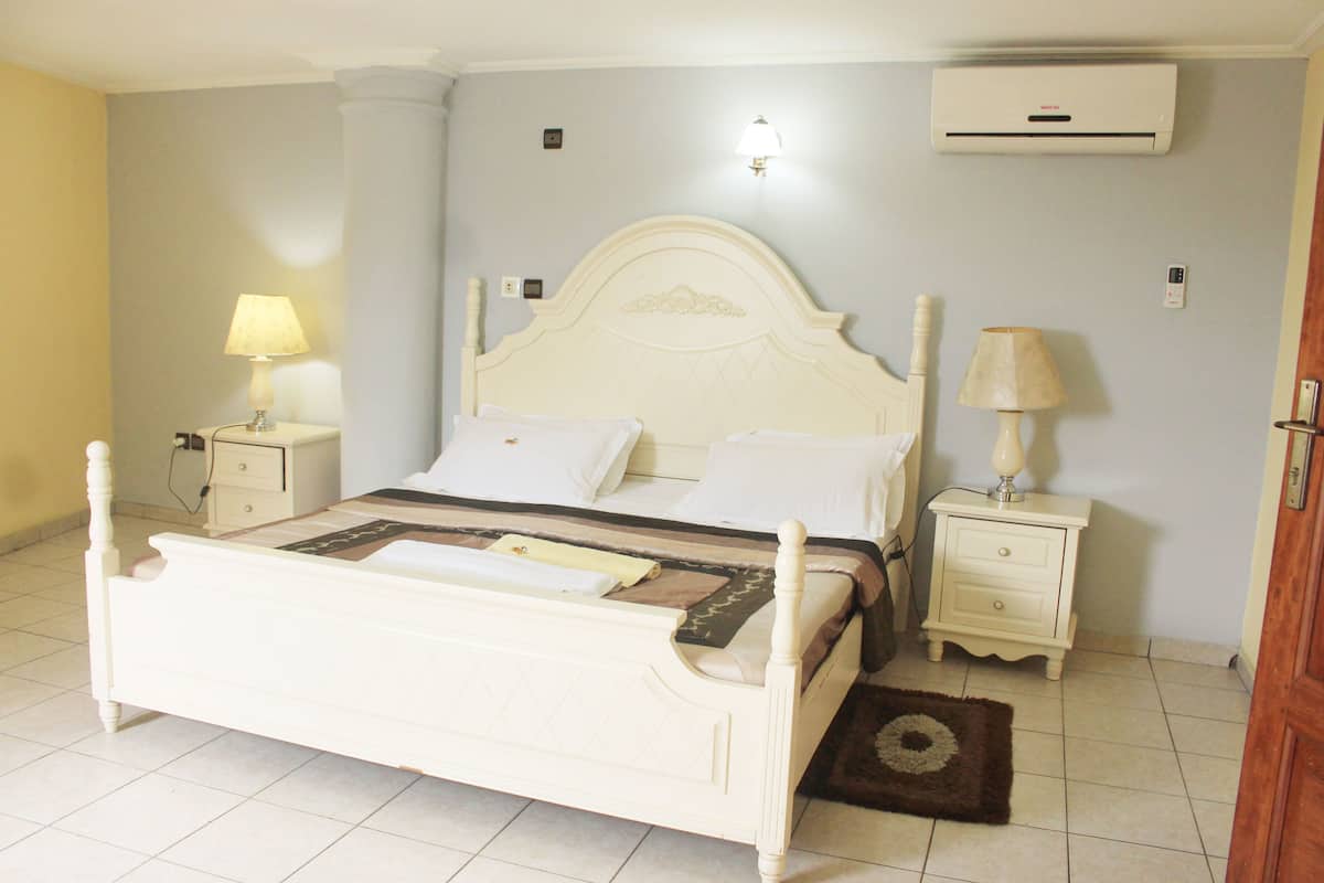 A bedroom with a white four-poster bed, two bedside tables with lamps, a wall-mounted air conditioner, and a small rug on the floor.