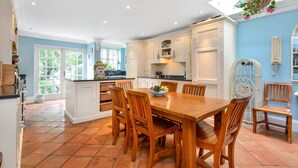 Une cuisine spacieuse avec une grande table à manger en bois, des meubles blancs et une vue sur l’extérieur grâce à de grandes fenêtres.