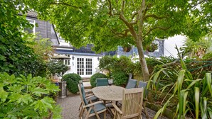 Un ensemble de table et de chaises en bois, placé sur une terrasse pavée, est entouré d’une végétation luxuriante. En arrière-plan, on aperçoit un bâtiment blanc avec un toit bleu.