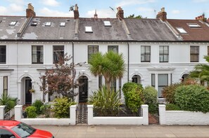 A row of white terraced houses with multiple windows, a gated entrance, and various plants in the front yard.