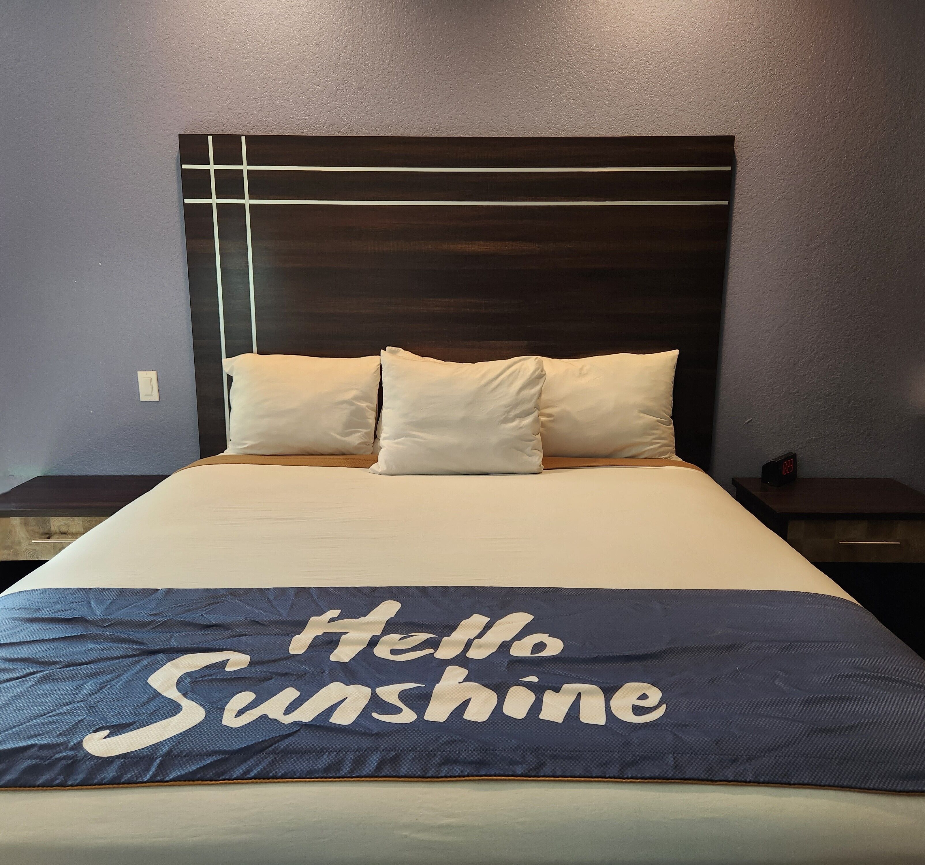 A bed with a headboard, two bedside tables, and a bedspread with "Hello Sunshine" text.