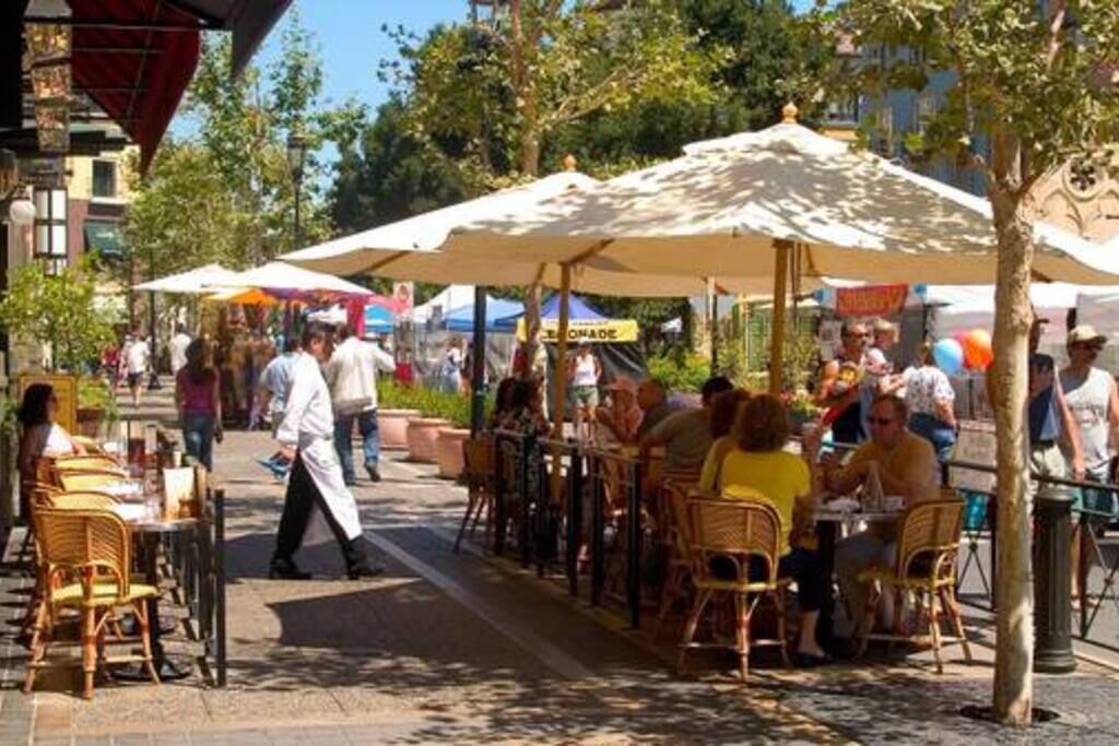 Ein Straßencafé mit Tischen und Stühlen unter Sonnenschirmen, umgeben von Bäumen und Gebäuden.