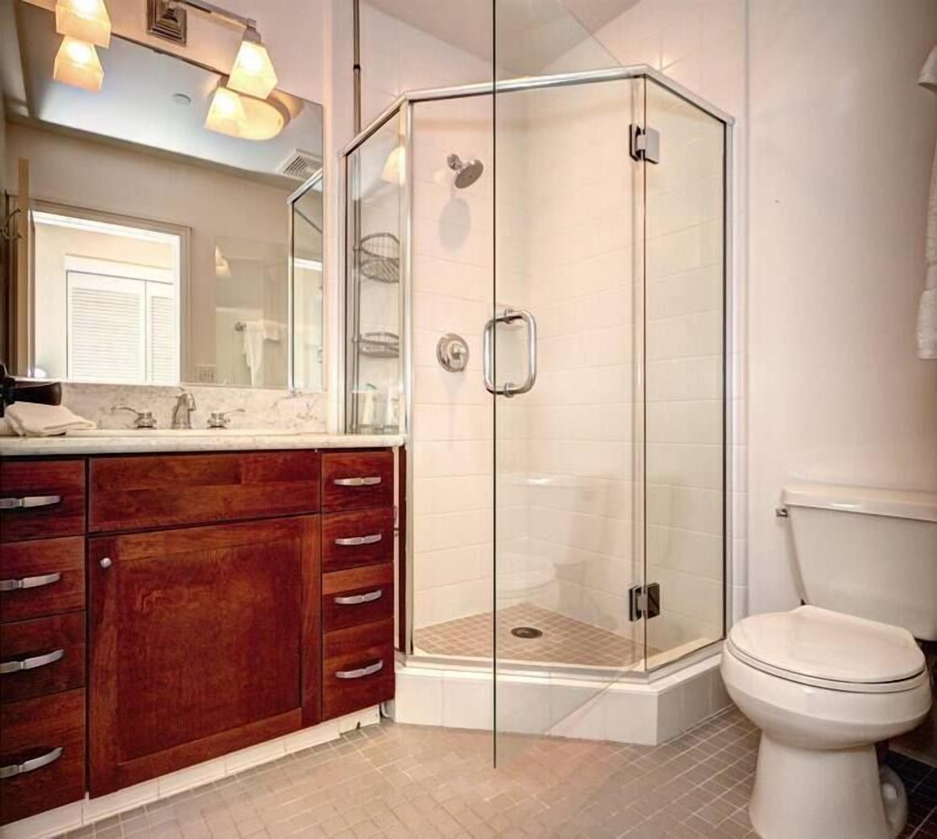 A modern bathroom with a glass-enclosed shower, a wooden vanity with multiple drawers, a white toilet, and a large mirror above the vanity.