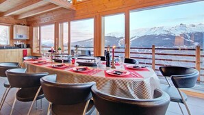 A dining area with a long table set for a meal, large windows with a mountain view, and wooden walls. - Chalet, wifi, ski-in/ski-out & foot of the slopes, sauna, terrace, balcony, fireplace or stove (Les Collons)