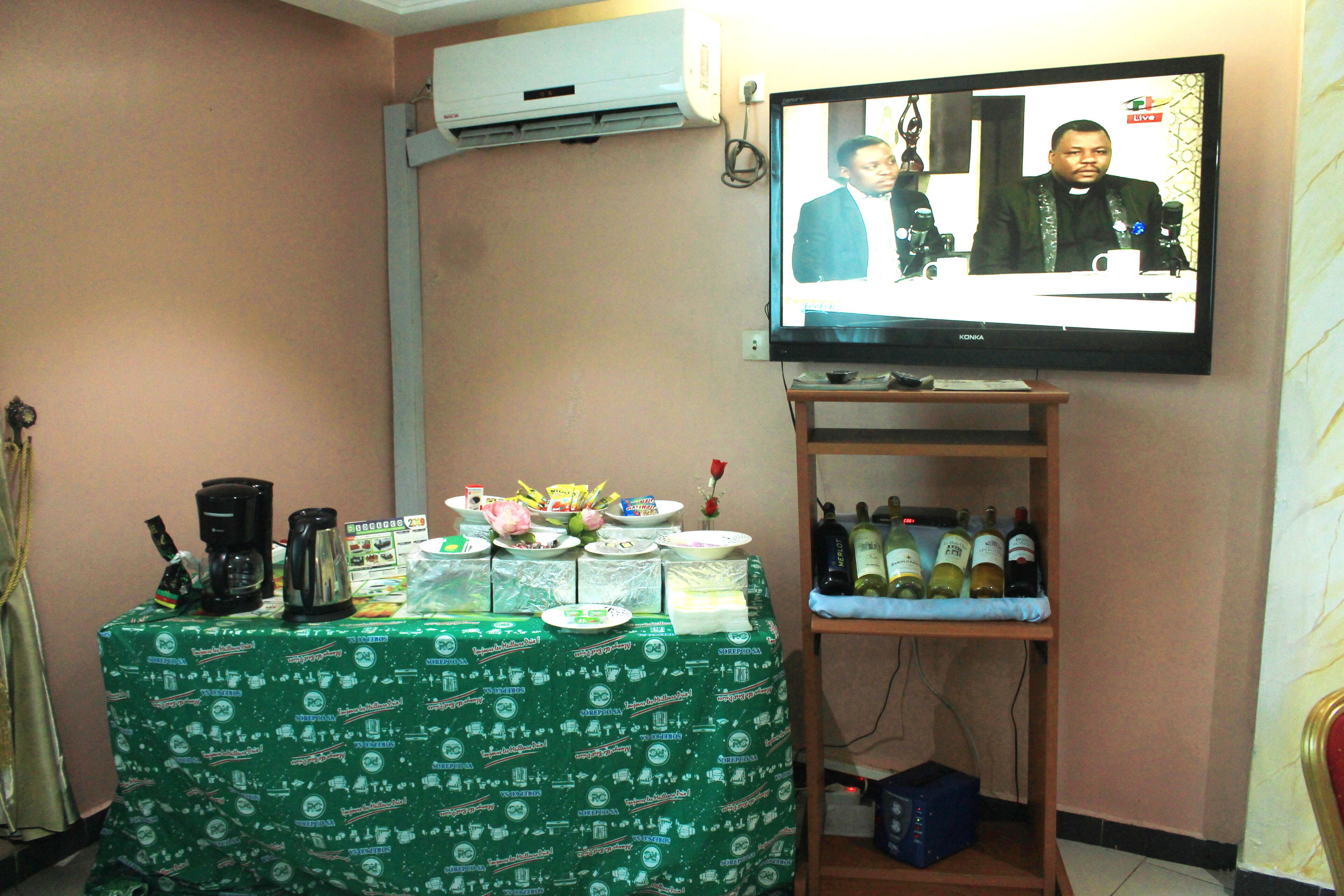 A room with a television displaying a live broadcast, a table with a green tablecloth, and a wine shelf.