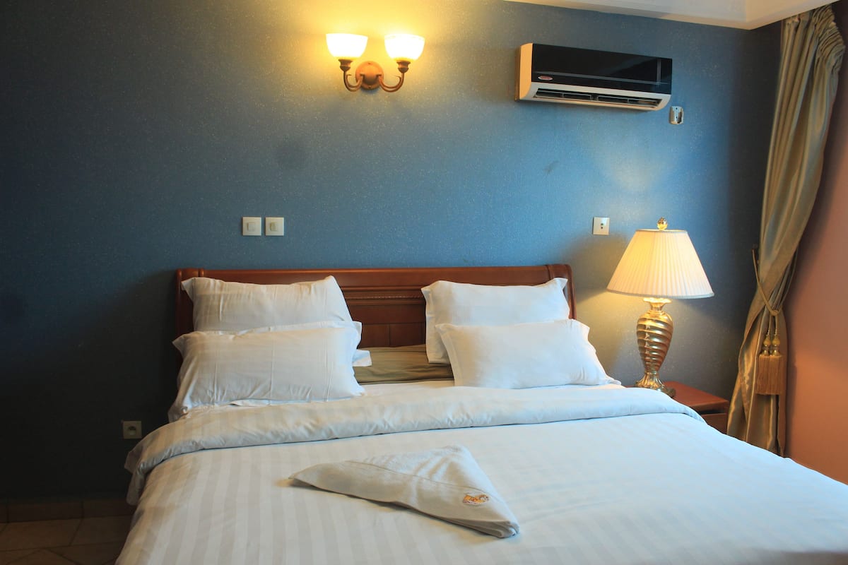 A hotel room with a bed, two bedside lamps, and a wall-mounted air conditioner.