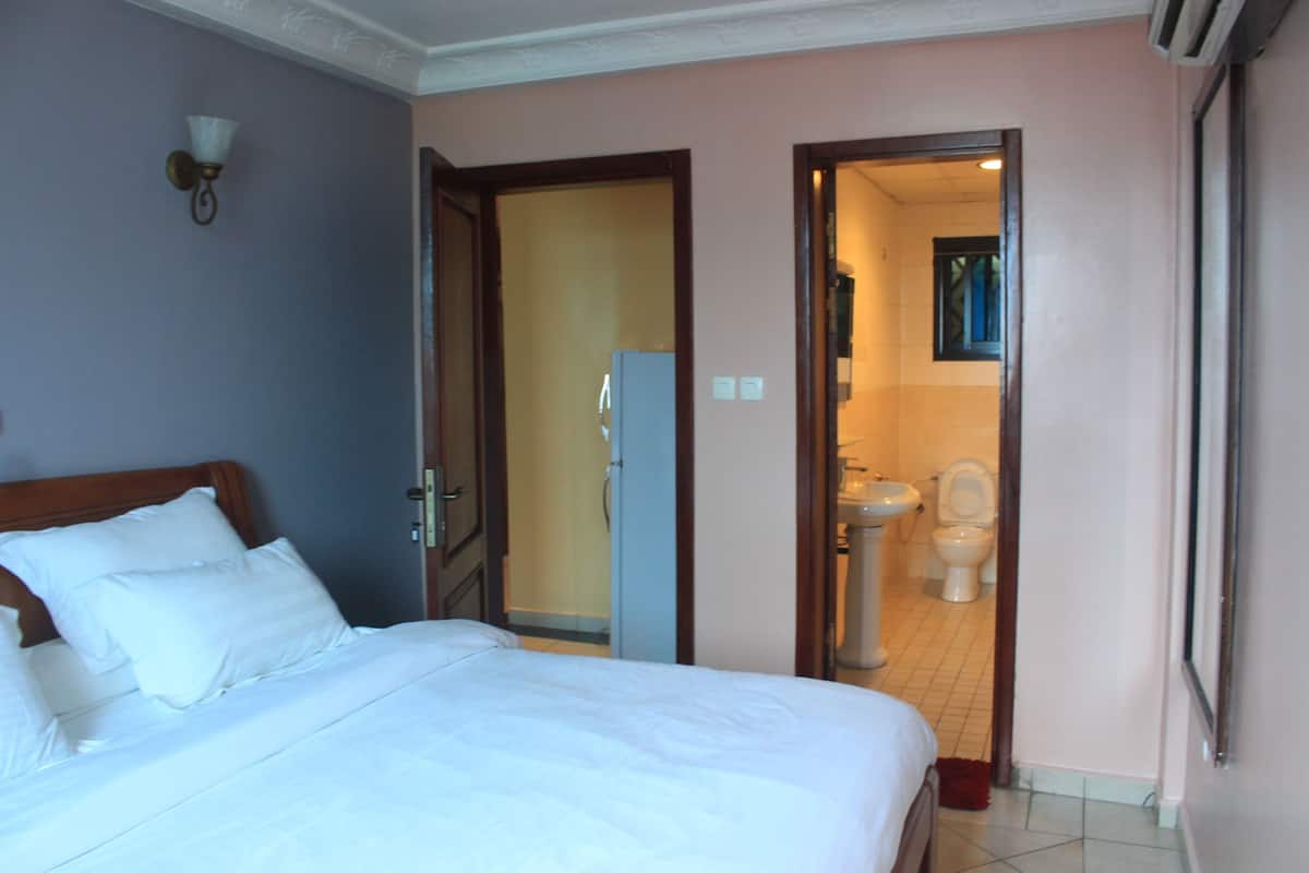 A hotel room with a bed, a nightstand, a wall-mounted light fixture, and an open door leading to a bathroom with a toilet and sink.