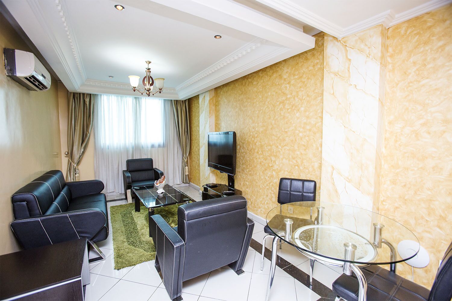 A modern living room with a sofa, armchairs, a glass coffee table, a TV, and a small dining table.