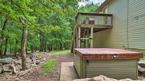 A wooden deck with a metal roof attached to a building, surrounded by a forested area with rocks and a concrete path. - Deck, Hot Tub & Game Room: Lake Harmony Home (Lake Harmony)
