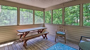 A covered outdoor area with a wooden picnic table and benches, large windows, and a chair with a cushion. - Deck, Hot Tub & Game Room: Lake Harmony Home (Lake Harmony)