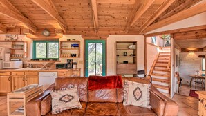 Un interior de cabaña acogedor con techo de madera, un sofá de cuero con cojines decorativos, una zona de cocina con armarios de madera y una escalera que sube a otro nivel.