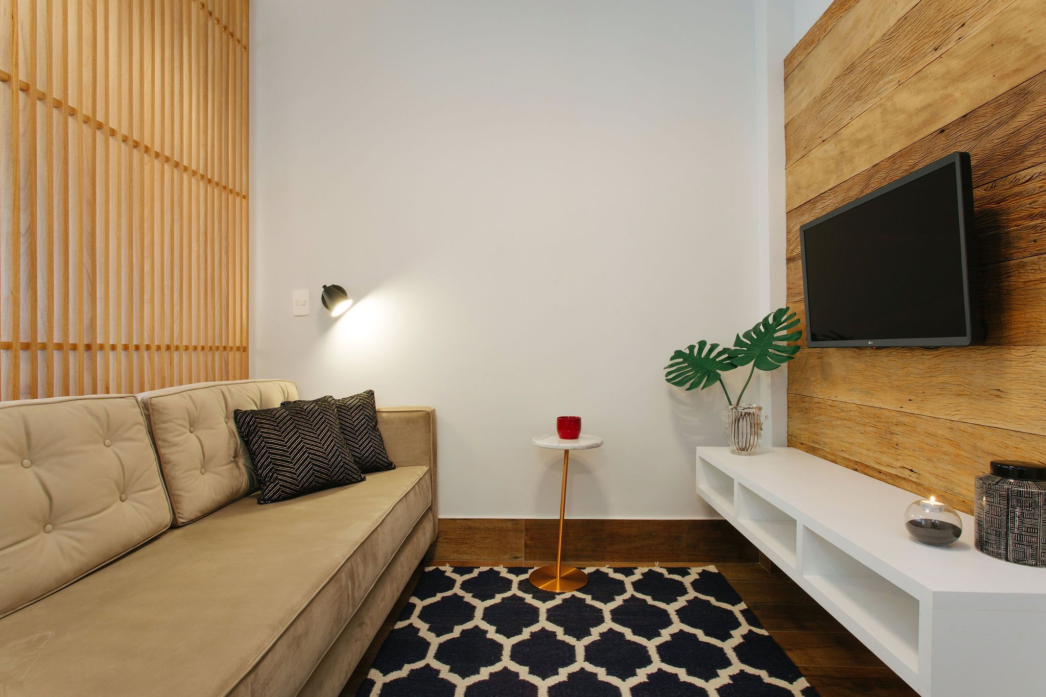 A modern living room with a beige sofa, a small round table with a red cup, a large flat-screen TV mounted on a wooden wall, and a decorative plant.