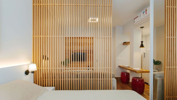Moderni hotellihuone, jossa on sänky, yöpöytä, puinen väliseinä, työpöytä tuolilla sekä pieni oleskelualue jakkaroilla.