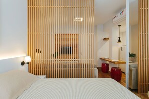 Une chambre d’hôtel moderne comprenant un lit, une table de chevet, une cloison en bois, un bureau avec une chaise et un petit coin salon avec des tabourets.
