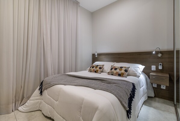 Un lit bien fait, avec une tête de lit, une table de chevet avec une lampe et un panneau de commande fixé au mur.