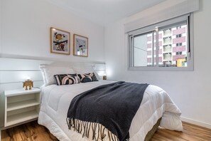 Quarto com cama, mesas de cabeceira, janela com vista para prédios e duas fotos emolduradas na parede.