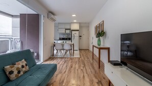 Un salon moderne avec un canapé vert, un téléviseur à écran plat et un coin repas, la cuisine étant visible au fond.