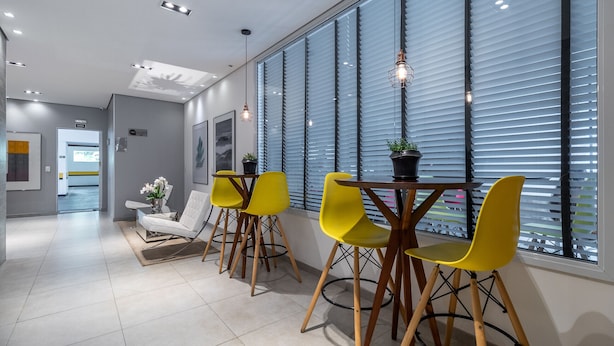 Un espace repas moderne avec des chaises jaunes, une table ronde et une vue dégagée grâce à de grandes fenêtres équipées de stores.